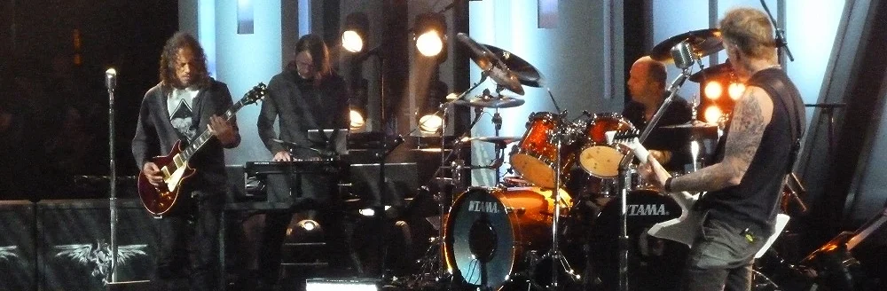On a television stage: Kirk Hammett on guitar, Sarth Calhoun at his keyboard and laptop, Lars Ulrich at the drums, James Hetfield on guitar.