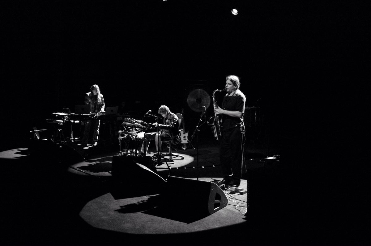 Metal Machine Trio on the Opera House stage, in black and white: Sarth Calhoun stands at laptops on the left, Lou Reed sits at his table of effects in the centre, and Ulrich Krieger plays saxophone in a spotlight on the right. Left to right: Sarth Calhoun, Lou Reed, Ulrich Krieger.