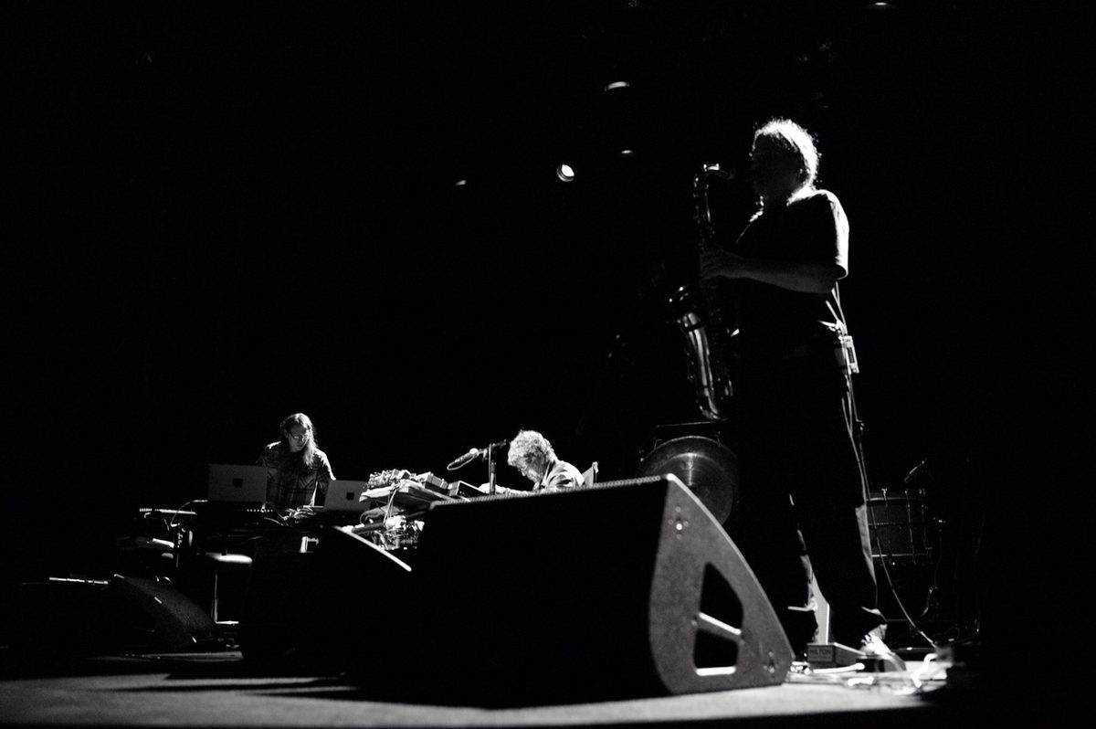 A low angle from the stage floor: Sarth Calhoun and Lou Reed are lit at their tables on the left and in the centre, and Ulrich Krieger’s silhouette with saxophone looms on the right behind a monitor wedge. Left to right: Sarth Calhoun, Lou Reed, Ulrich Krieger.