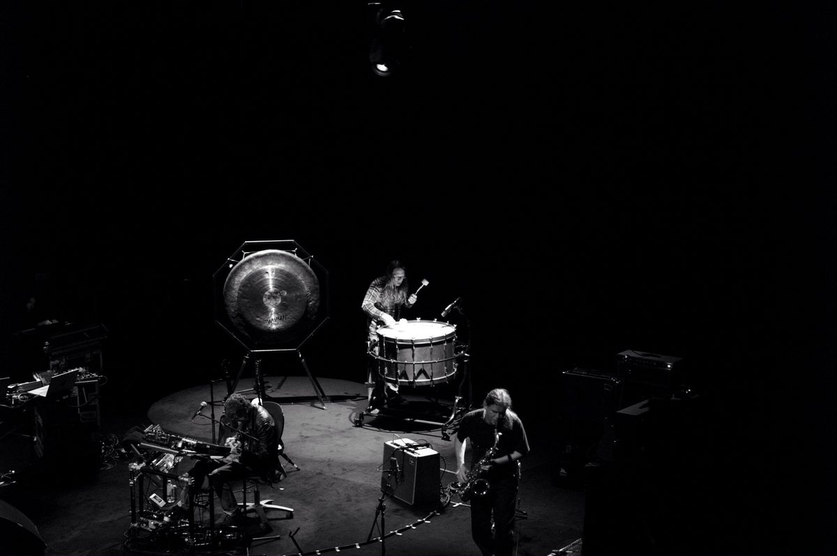 Seen from above: Lou Reed sits at his effects on the left, Sarth Calhoun strikes a large bass drum beside a gong at the back of the stage, and Ulrich Krieger plays saxophone at the front right. Left to right: Lou Reed, Sarth Calhoun, Ulrich Krieger.