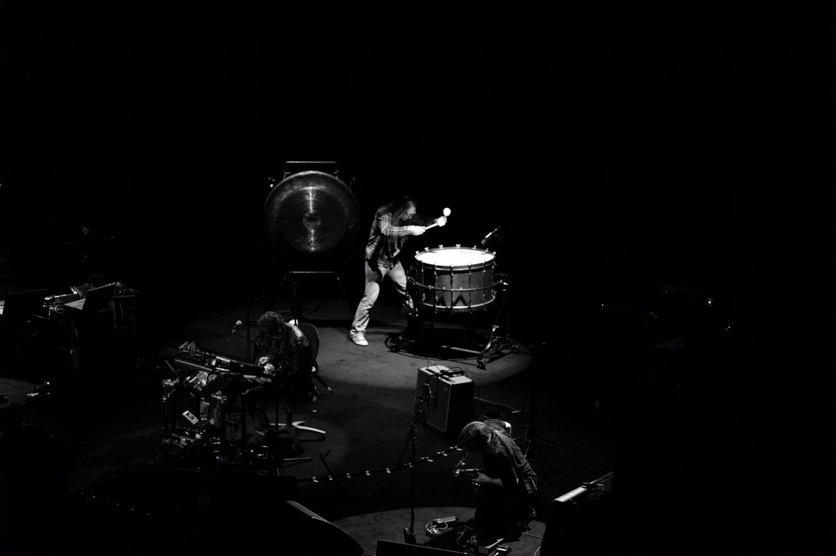 Seen from above: Lou Reed bends over his effects on the left, Sarth Calhoun swings a mallet at the bass drum beside the gong, and a player crouches over gear in the foreground. Left to right: Lou Reed, Sarth Calhoun.