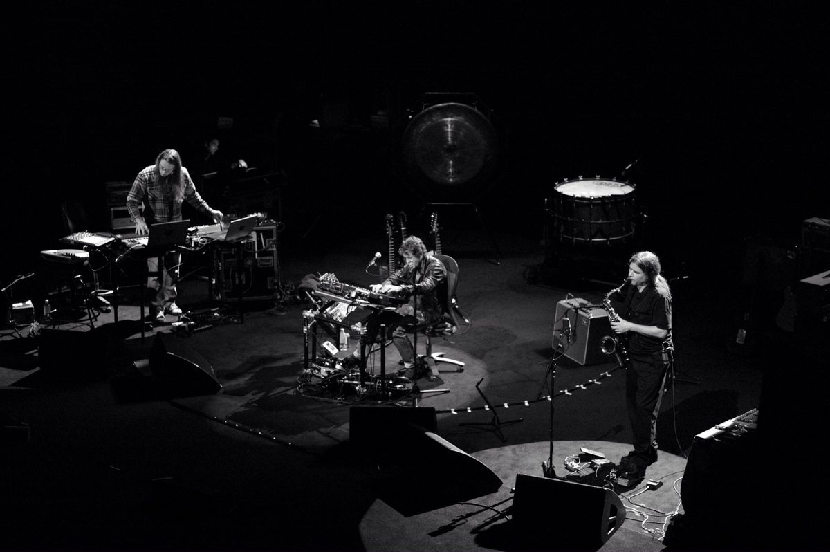 A wide view from above: Sarth Calhoun at laptops on the left, Lou Reed seated in the centre with guitars on stands behind him, Ulrich Krieger with saxophone on the right, and the gong and bass drum at the back. Left to right: Sarth Calhoun, Lou Reed, Ulrich Krieger.