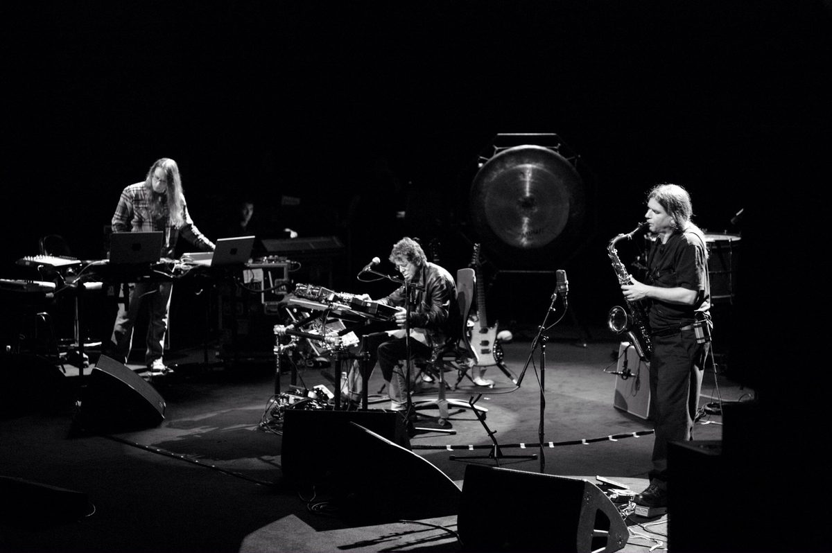 Sarth Calhoun at laptops on the left, Lou Reed seated at his effects in the centre with a guitar on a stand beside him, and Ulrich Krieger playing saxophone on the right, the gong behind them. Left to right: Sarth Calhoun, Lou Reed, Ulrich Krieger.