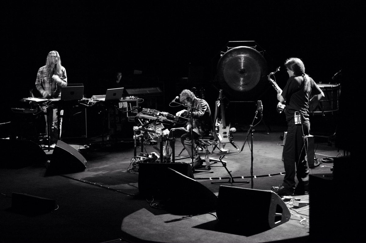 Sarth Calhoun stands at laptops on the left, Lou Reed works his effects in the centre, and Ulrich Krieger, seen from behind, turns toward him holding his saxophone, with the gong and bass drum at the back. Left to right: Sarth Calhoun, Lou Reed, Ulrich Krieger.