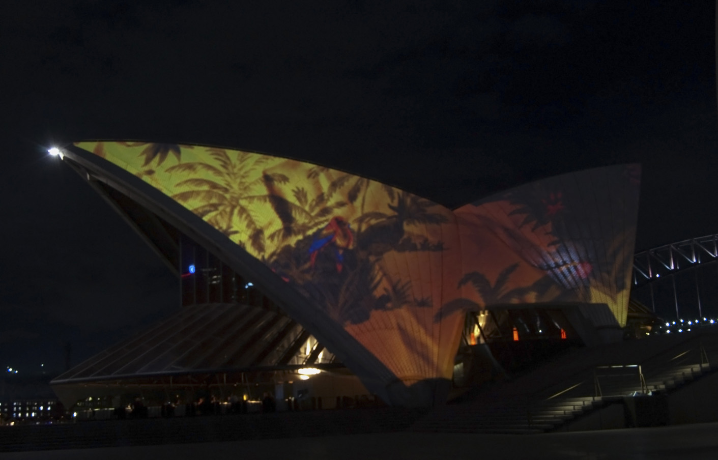 The sails of the Sydney Opera House at night, lit with a projection of palms and a parrot.